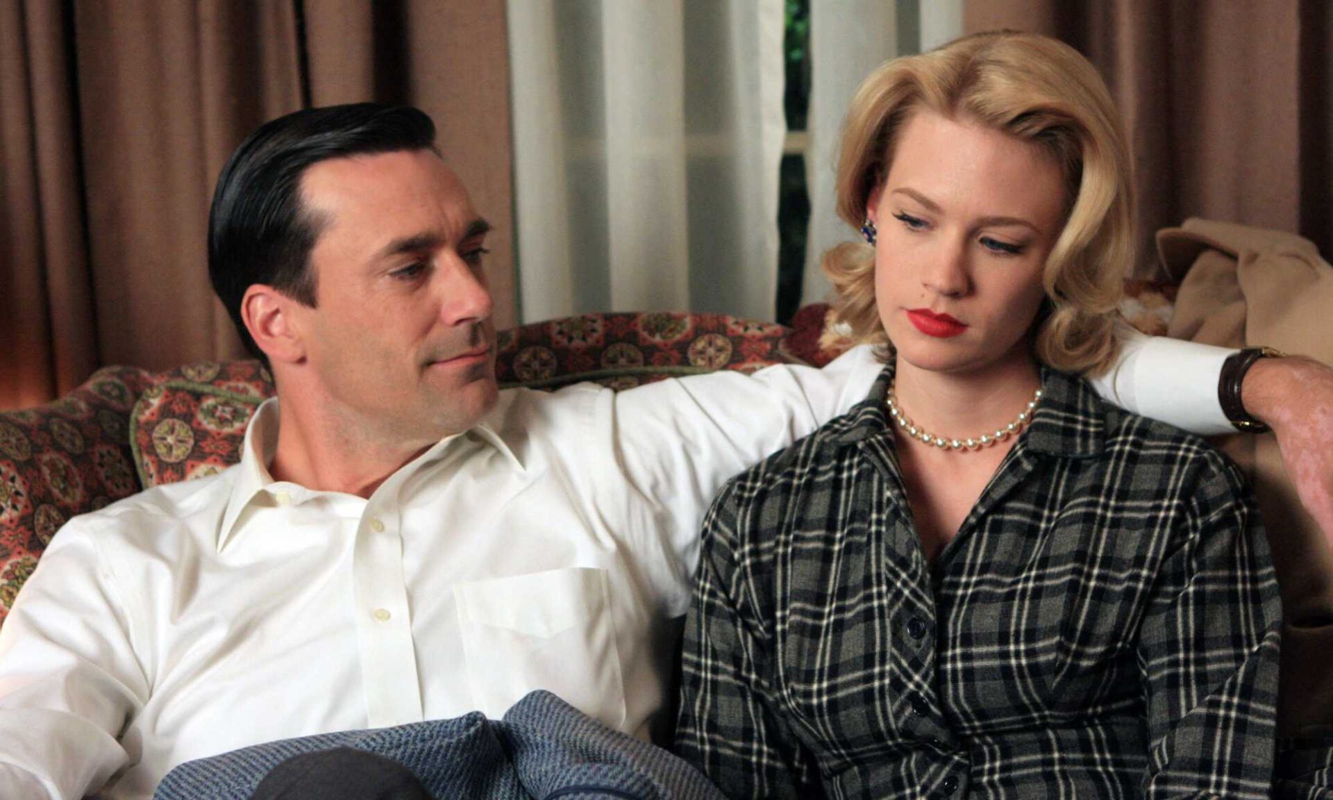 Still from the TV show Mad Men with Don Drapper sitting on a couch with his around around wife Betty to illustrate the idea of power dynamics and infidelity.